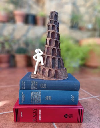 Una versión esculpida en barro de la Torre de Babel, símbolo de la traducción, se sostiene sobre una pequeña pila de tres gruesos diccionarios de términos jurídico-económicos. Este grupo está colocado en primer plano sobre el suelo de un patio con un fondo difuminado de plantas en macetas.